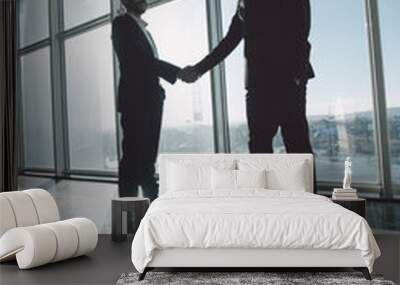 Two young businessmen are shaking hands with each other standing against panoramic windows. Wall mural